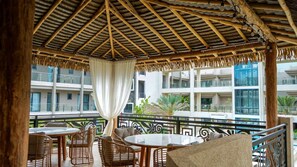 An outdoor seating area with wicker furniture, a thatched roof, and a view of a modern building. - Sunset Balconies | 5 minutes to Eagle Beach (Noord)