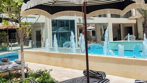 Zona de piscina con sillones de playa y sombrillas, un edificio decorativo con un gran diseño floral, y un cielo azul despejado.
