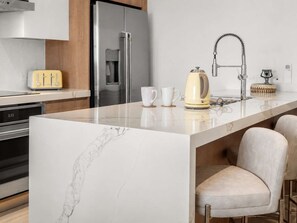 Une cuisine moderne dotée d’un plan de travail en marbre, d’appareils électroménagers en acier inoxydable, d’une bouilloire et d’un tabouret de bar.