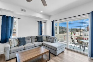 A modern living room with a sectional sofa, a coffee table, and a large sliding glass door leading to an outdoor patio area. - Luxury Jersey Shore Retreat Rooftop Oasis & Beach (Seaside Heights)