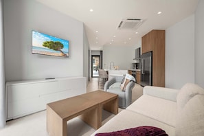 A modern living room with a flat-screen TV, a wooden coffee table, a grey sofa, and a kitchen area in the background. - 5-Min Drive to Eagle Beach|Large Patio|Pool&Jacuzzi (Noord)