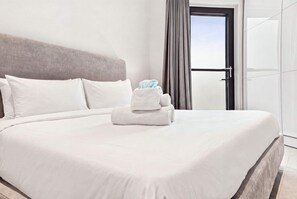 Ein ordentlich bezogenes Bett mit weißen Leinen und einem gefalteten Handtuch, ein grauer Kopfteil und ein Blick ins Freie durch eine Tür mit Vorhängen.