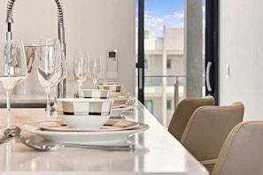 A dining table set with plates, glasses, and cutlery, with a view of buildings through large windows. - Top-Floor 1BR | Resort Pool | Close to Eagle Beach (Noord)