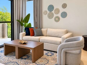 Un salon moderne avec un canapé, une table basse et un fauteuil. Il y a une grande fenêtre avec des rideaux, une plante et des œuvres d’art murales.