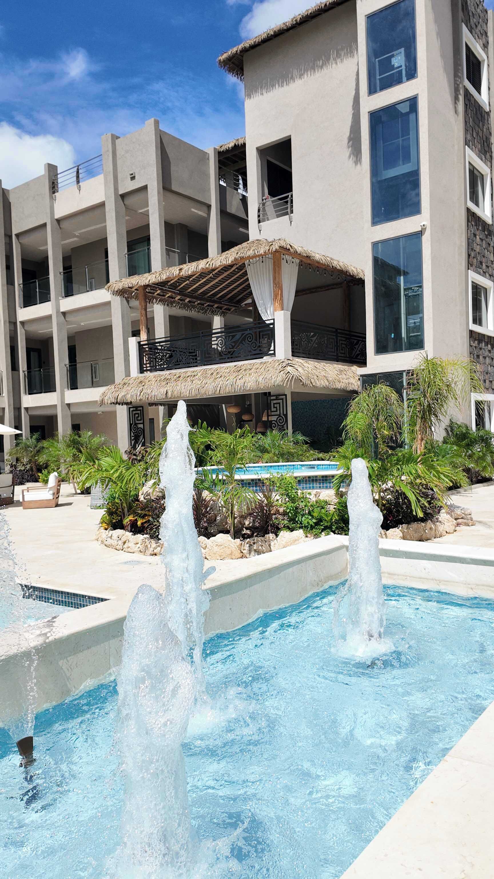A swimming pool with water fountains, surrounded by tropical plants and a building with balconies.