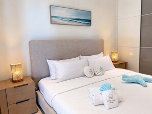 A neatly made bed with white bedding and two decorative pillows, a bedside lamp, and a wall-mounted painting. - Sunrise & Sunset | Corner 1BR | Near Eagle Beach (Noord)