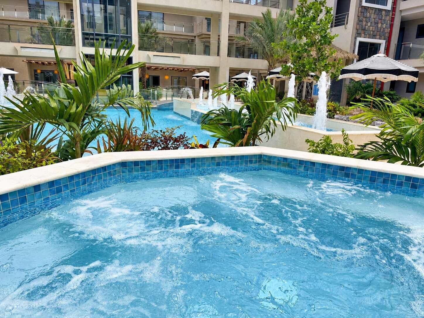 Zona de piscina hotelera con varios niveles de balcones, palmeras y una fuente.