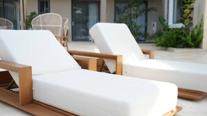 Zwei weiße Lounge-Stühle mit Holzgestell, platziert auf einer Terrasse mit Blick auf ein Gebäude und Grünflächen im Hintergrund.