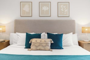 Ein ordentlich bezogenes Bett mit Kopfteil, Kissen in Weiß und Türkis sowie ein beiges Zierkissen und eine kleine beige Tasche auf dem Bett. Darüber hängen drei gerahmte Wandbilder.