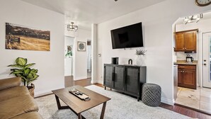 Ein Wohnzimmer mit Sofa, Couchtisch, einem an der Wand montierten Fernseher und einer Küchenzeile mit Schränken und einer Kaffeemaschine.