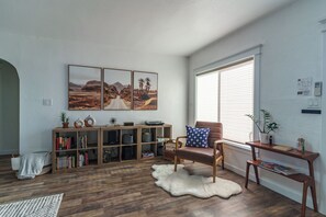 Un salón con una estantería de madera, un sillón de cuero, un manto de piel sintética y una ventana con persianas.