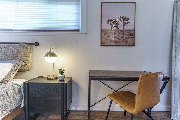 A bedroom with a bed, bedside table with a lamp, a desk, a chair, and a framed picture on the wall.