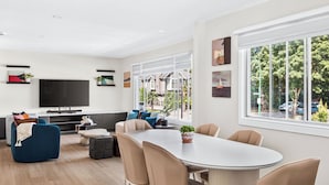 Un salón moderno con televisor de pantalla plana, un sofá, una mesa de comedor con sillas y un amplio ventanal con vistas al exterior.