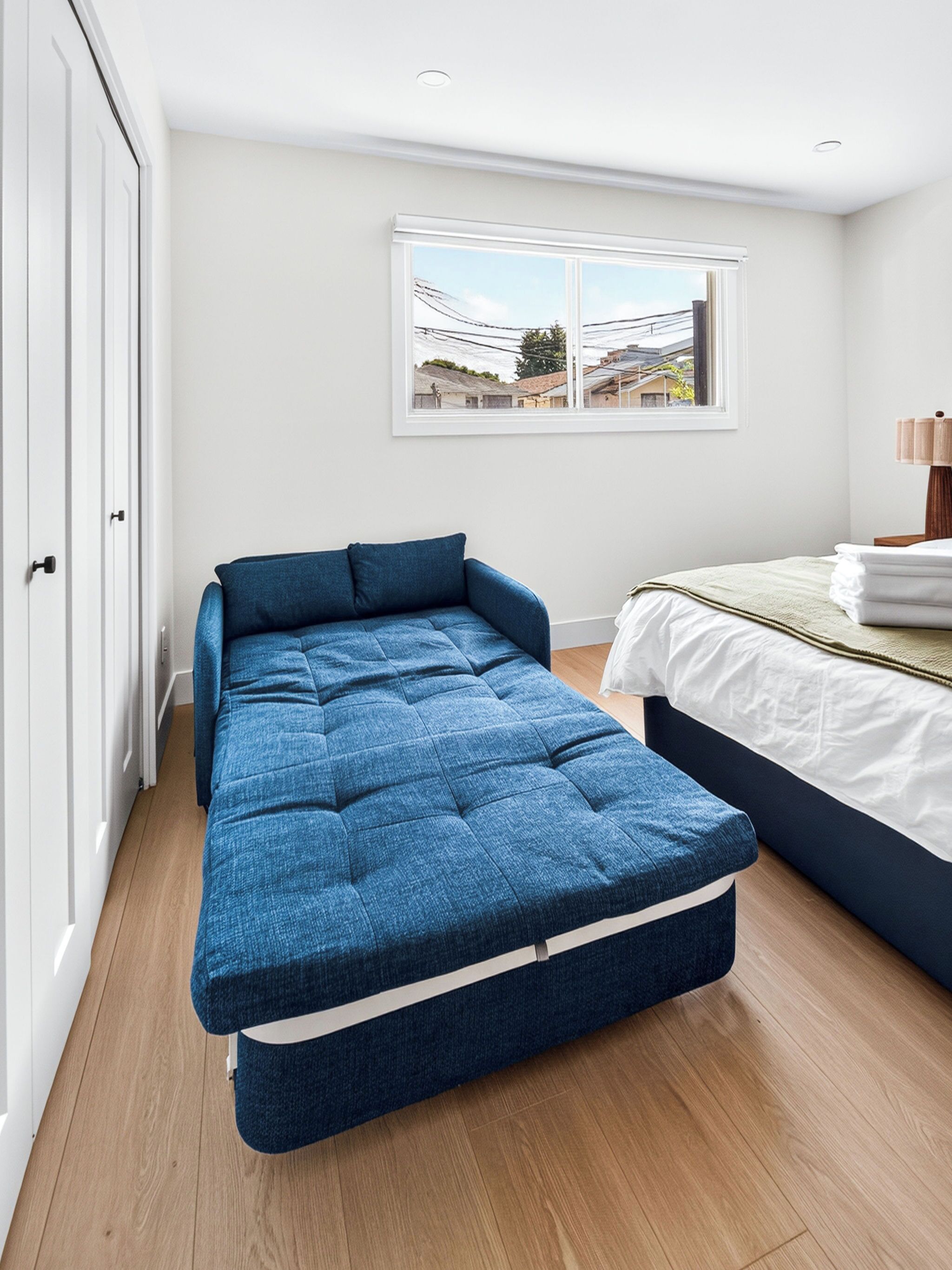 A room with a bed, a blue sofa bed, a window with a view of buildings, and a door.