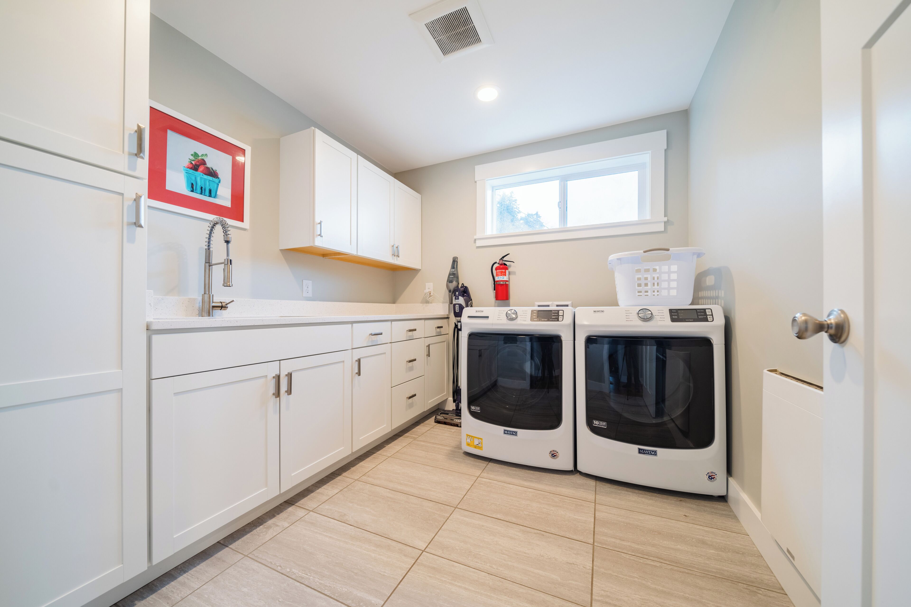 A laundry room with two Maytag washing machines, a fire extinguisher, and a window above the machines.