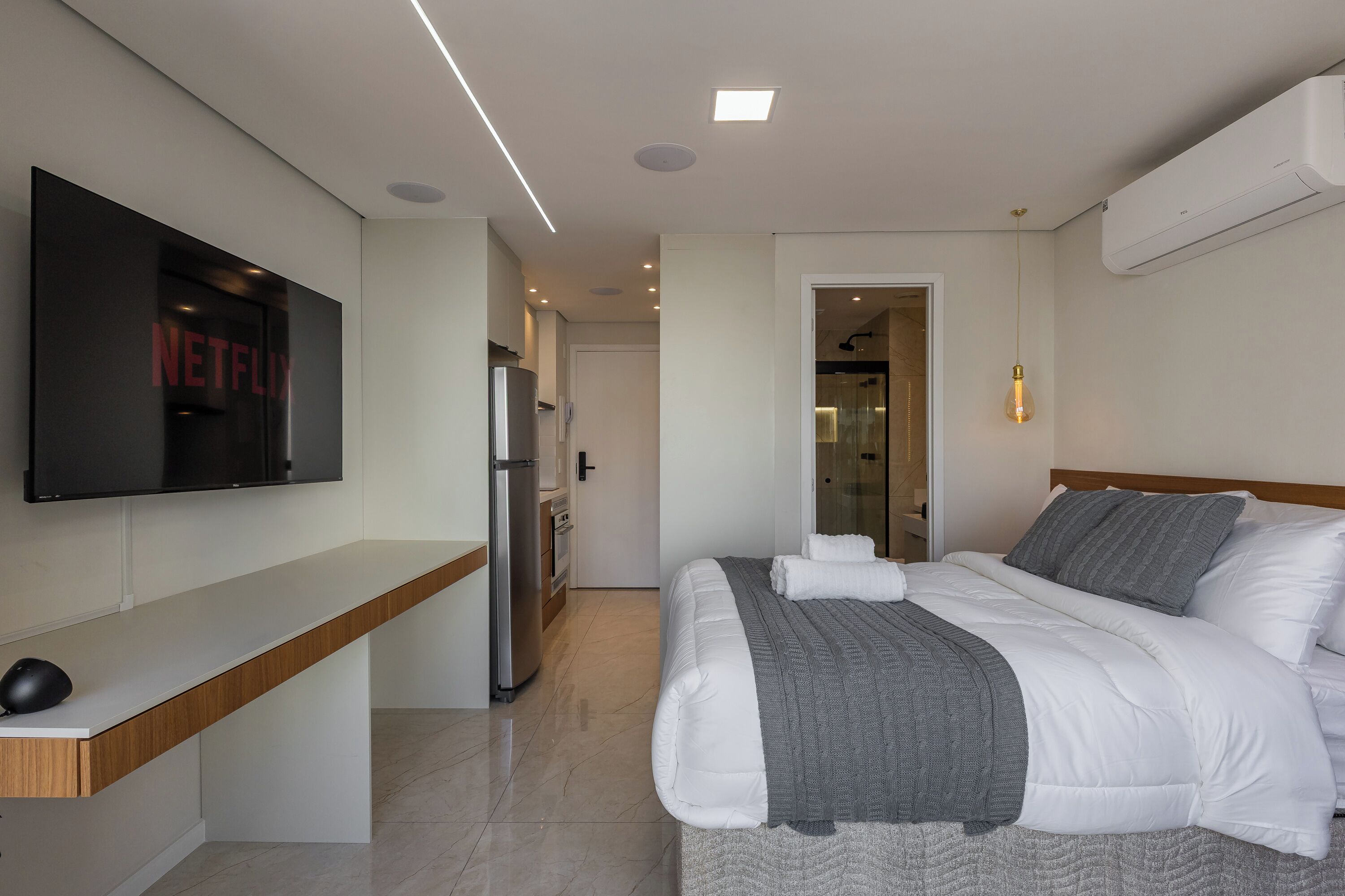 Quarto de hotel moderno com televisão de tela plana, cama com roupa de cama branca e cinza, mesa de cabeceira de madeira, mini-geladeira embutida e área de chuveiro.