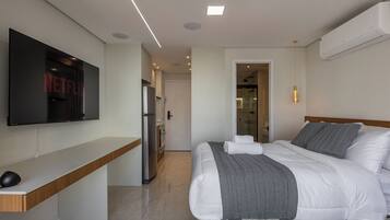 Quarto de hotel moderno com televisão de tela plana, cama com roupa de cama branca e cinza, mesa de cabeceira de madeira, mini-geladeira embutida e área de chuveiro.