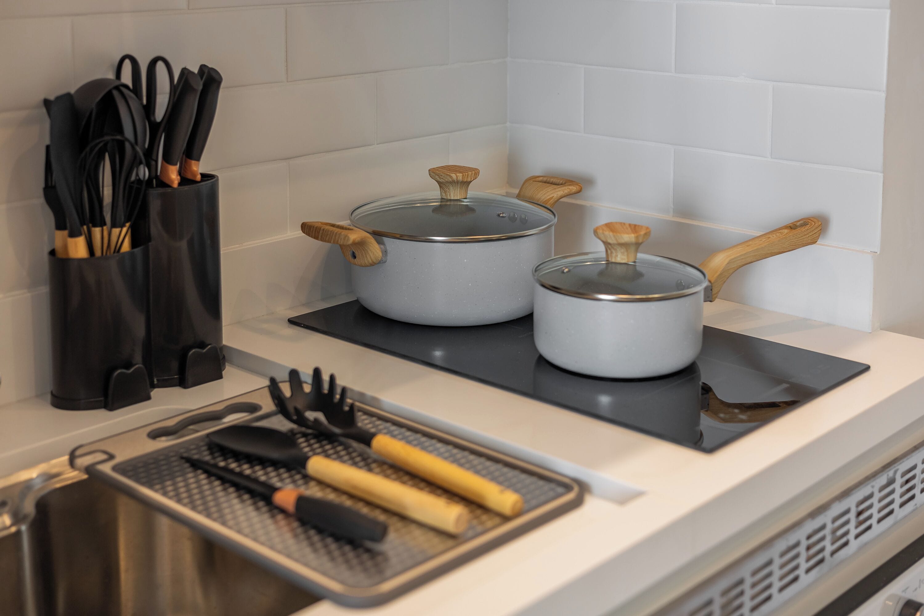 Uma cozinha com duas panelas em um fogão por indução preto, uma tábua de corte com utensílios, e um suporte preto para talheres com facas.
