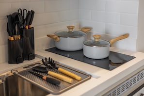 Uma cozinha com duas panelas em um fogão por indução preto, uma tábua de corte com utensílios, e um suporte preto para talheres com facas.