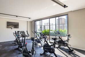 A gym with stationary bikes and a large window with a city view. - StayHome | Level Brooklin | Gym & Pool (São Paulo)
