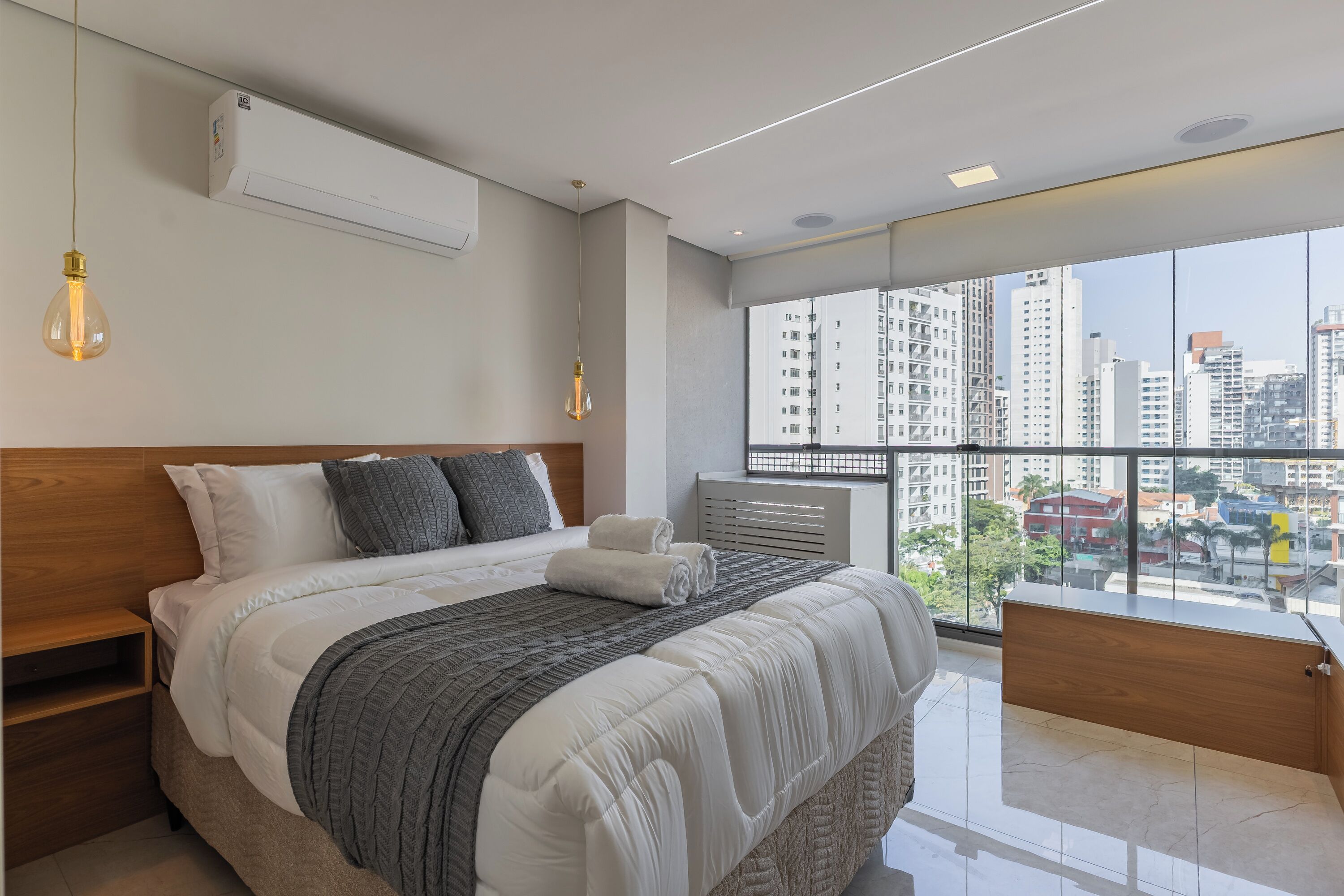Un dormitorio moderno con una cama grande, vistas de la ciudad a través de ventanales y un aire acondicionado integrado.
