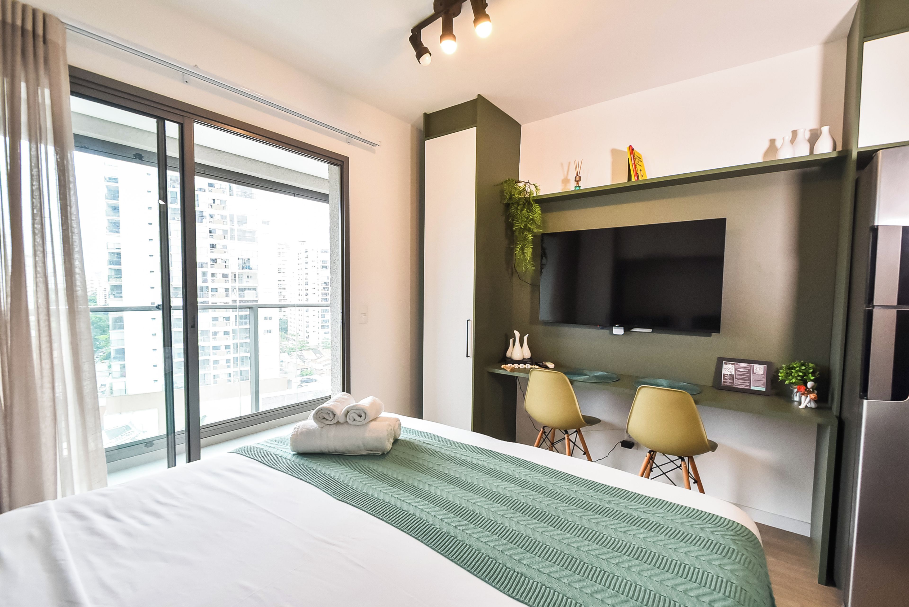Ein modernes Hotelzimmer mit einem großen Bett, einem Flachbildfernseher an einer dunklen Wand, einem kleinen Schreibtisch mit Stuhl und einem Fenster mit Blick auf hohe Gebäude.