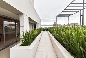 Um corredor com piso de azulejos brancos, ladeado por plantas verdes, que leva a uma entrada de prédio com porta de vidro.