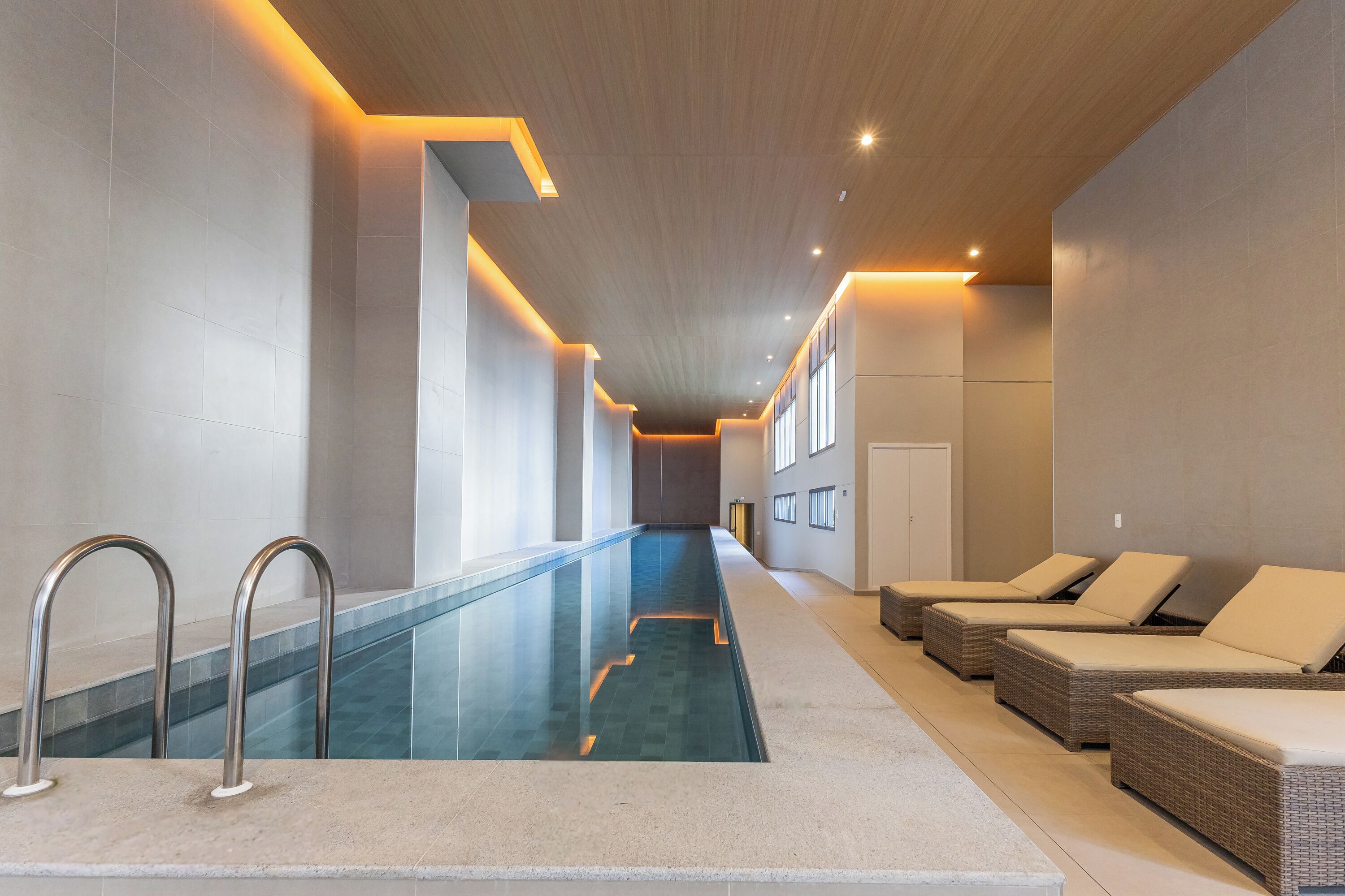 Piscina interior moderna com água cristalina, área de estar adjacente com cadeiras estofadas e um longo corredor com teto de madeira.