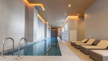 Piscina interior moderna com água cristalina, área de estar adjacente com cadeiras estofadas e um longo corredor com teto de madeira.