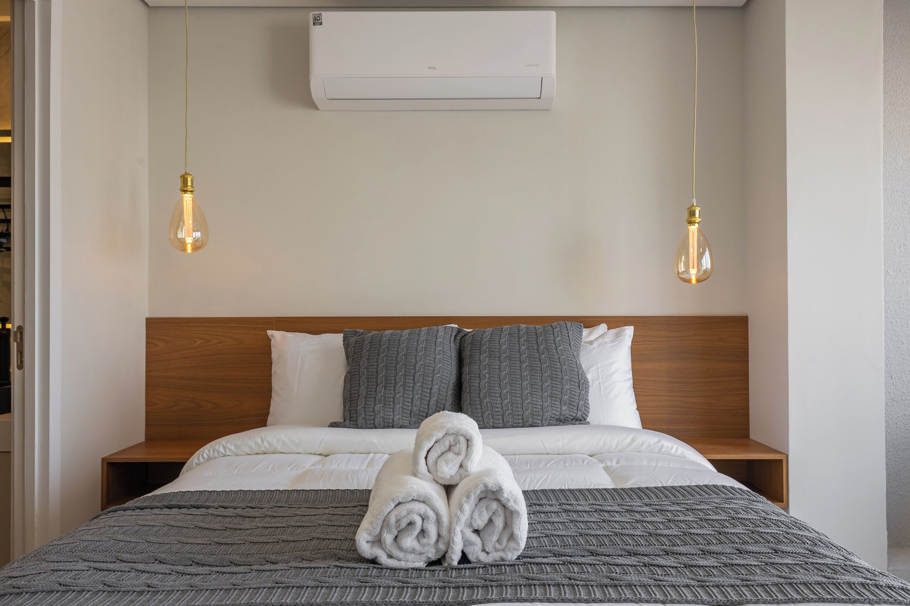 Une chambre d’hôtel avec un lit, une tête de lit en bois, une couverture tricotée grise, des serviettes blanches roulées, deux oreillers gris et un appareil de climatisation blanc.