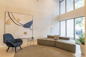 A modern living room with a sectional sofa, a blue armchair, a round rug, a floor lamp, and a large abstract painting on the wall. - StayHome | Level Brooklin | Gym & Pool (São Paulo)