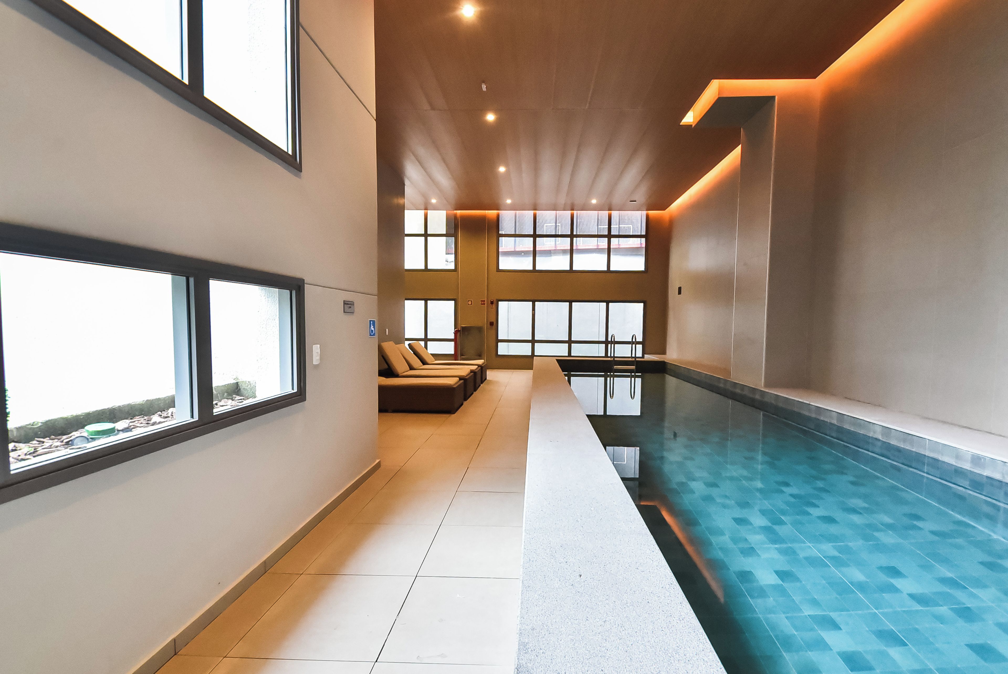 A modern indoor swimming pool with clear glass walls, a long bench seating area, and a view of the outside.