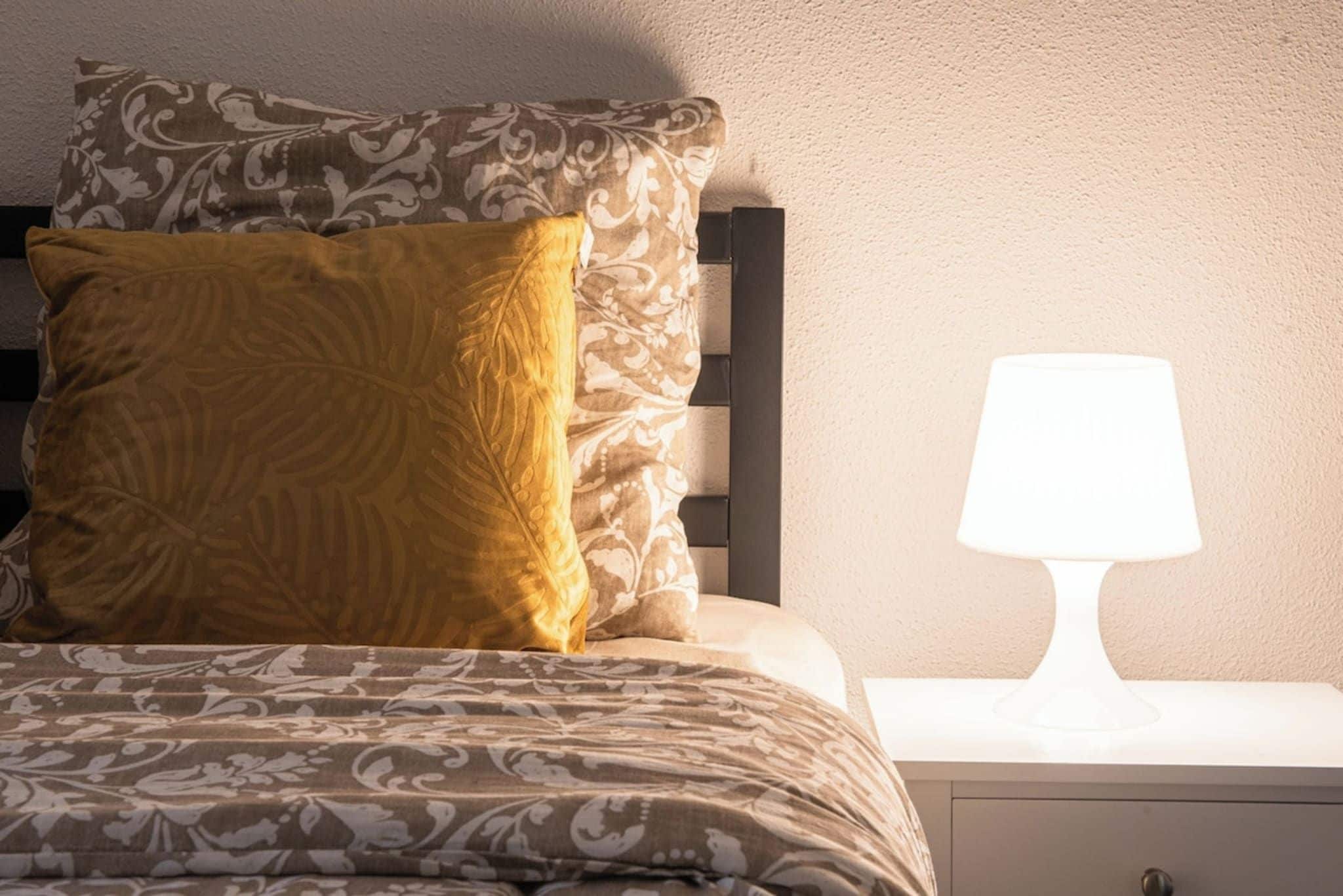 Een bed met beddengoed met een patroon en een enkel kussen, een nachtkastje met een lamp, en een effen muur.