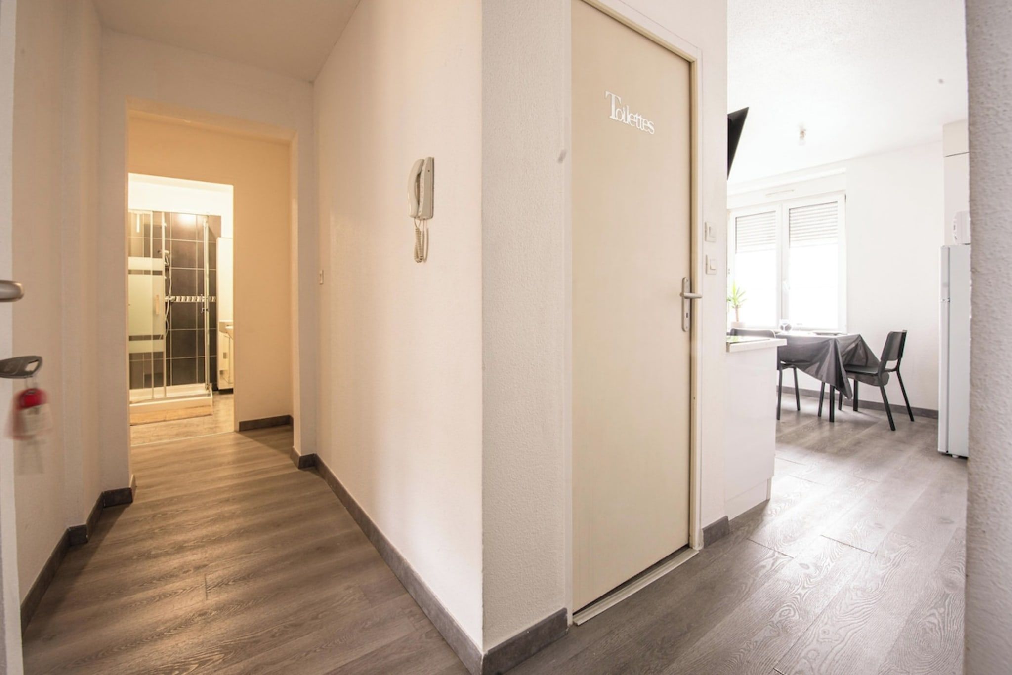 Un couloir mène à une pièce où se trouvent des toilettes, une porte, une table avec des chaises et une fenêtre avec des stores.