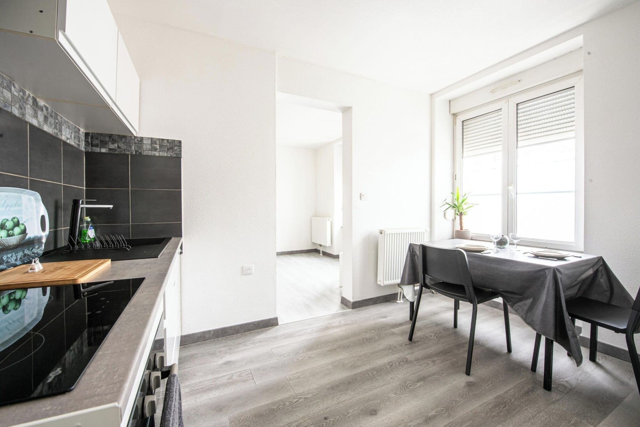 Eine moderne Küche mit Essbereich, ausgestattet mit einer schwarzen Arbeitsplatte, einem Esstisch mit Stühlen und einem Fenster mit Jalousien.