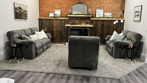 A living room with two grey sofas, a coffee table, a fireplace, and a brick wall. - Adorable 2-bedroom apartment with jetted Jacuzzi tub adjacent to Coffee Shop! (Brookfield)