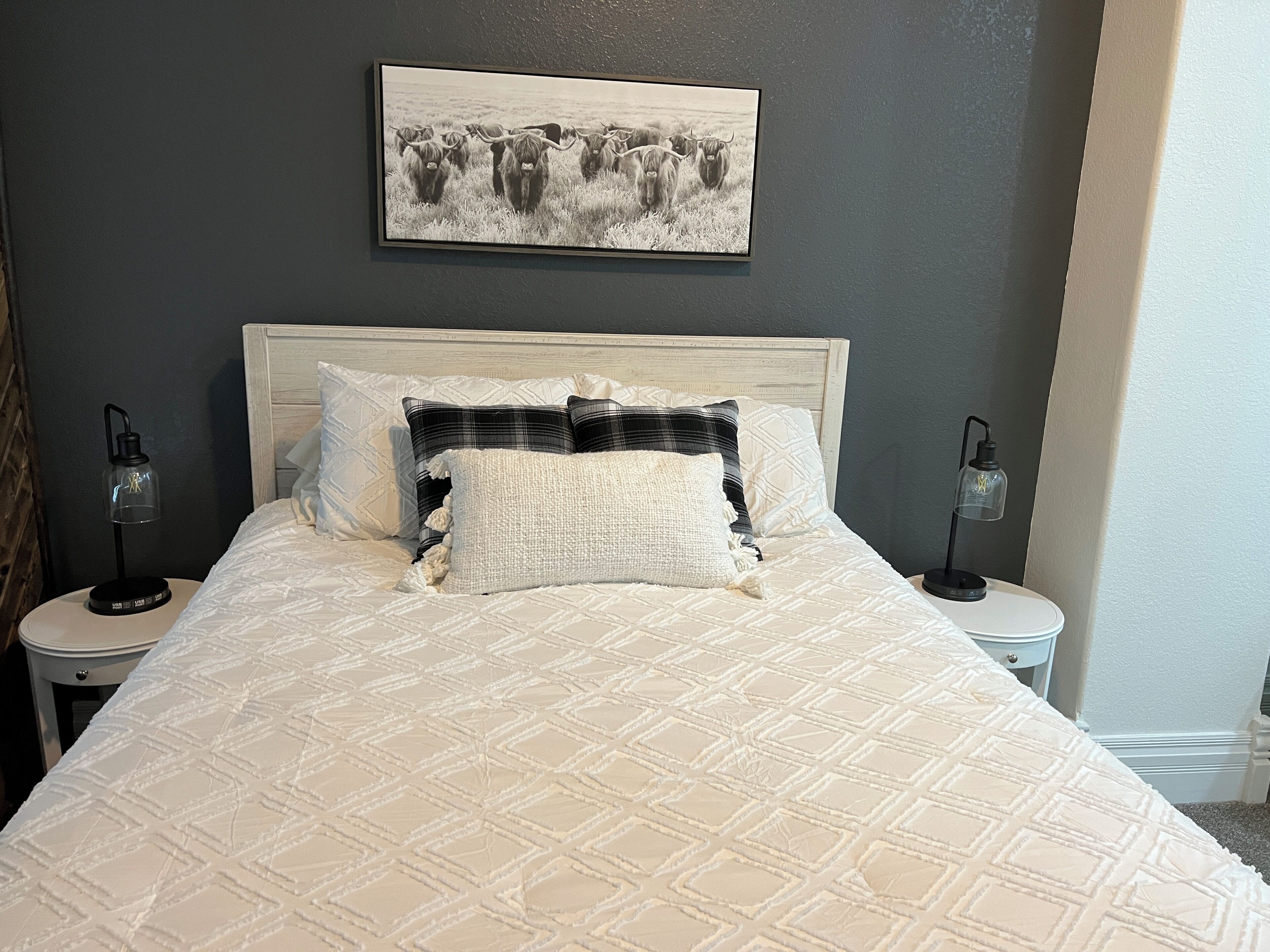 A bed with a quilted pattern, two bedside tables with lamps, a framed picture on the wall, and a dark-colored wall.
