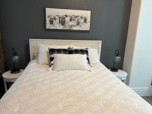 A bed with a quilted pattern, two bedside tables with lamps, a framed picture on the wall, and a dark-colored wall.
