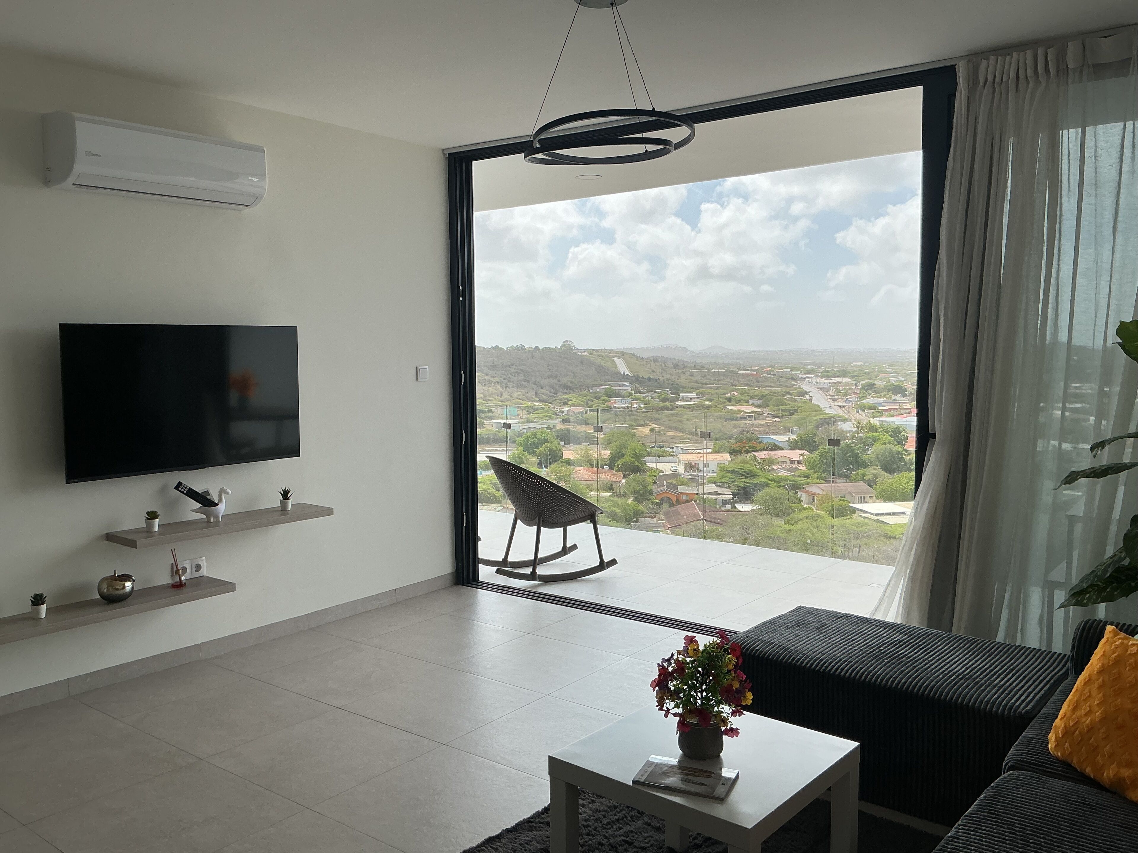 A modern living room with a flat-screen TV, a sofa, a coffee table with a vase, and a view of a cityscape through a large window.