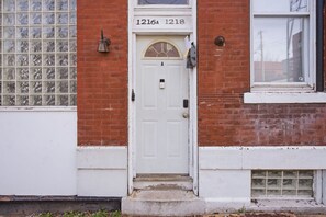 A white door with a small window above it, flanked by two brick walls, and a set of numbers '1216A 1218' above the door.