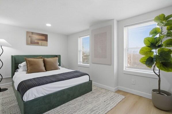 A modern bedroom with a green bed, white walls, a large window, and a potted plant.