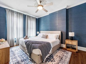 Une chambre à coucher avec un lit, des tables de chevet, un ventilateur de plafond et une fenêtre avec des rideaux.
