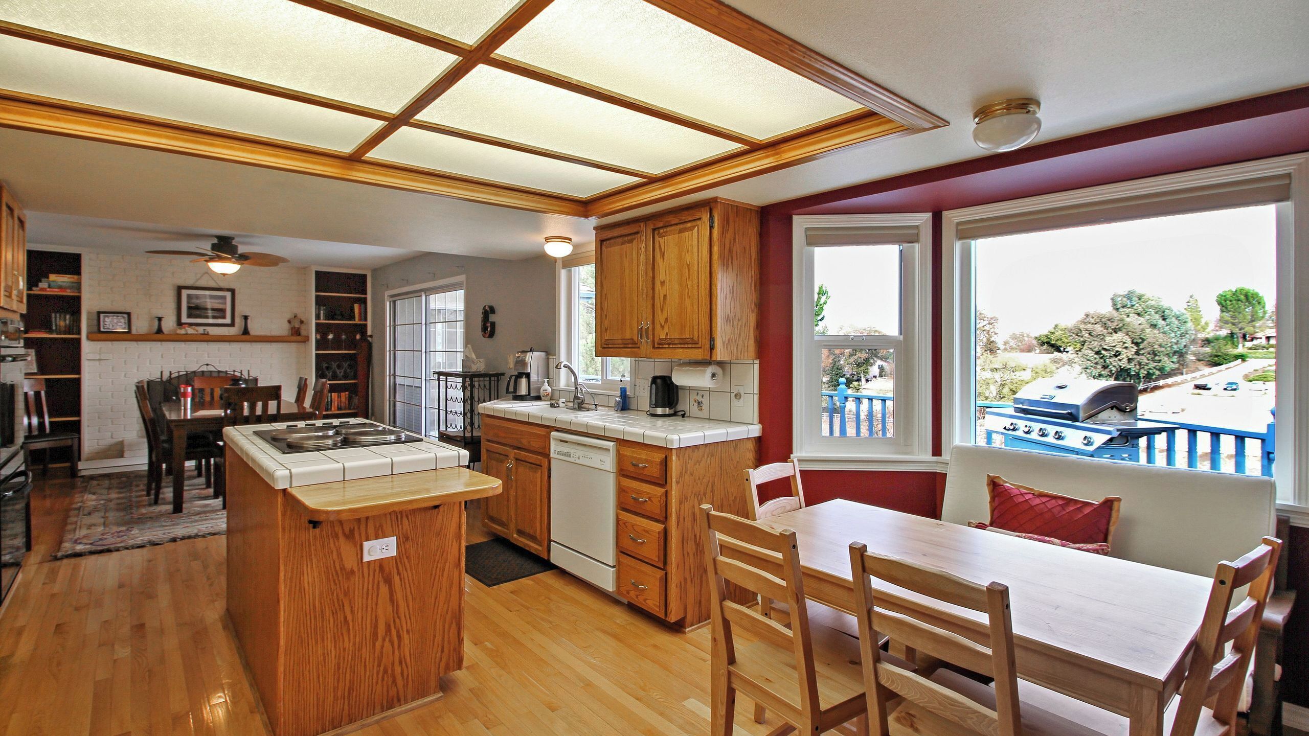 A kitchen with a central island, wooden cabinets, a dining area with a table and chairs, and a view of a blue deck and barbecue grill outside.