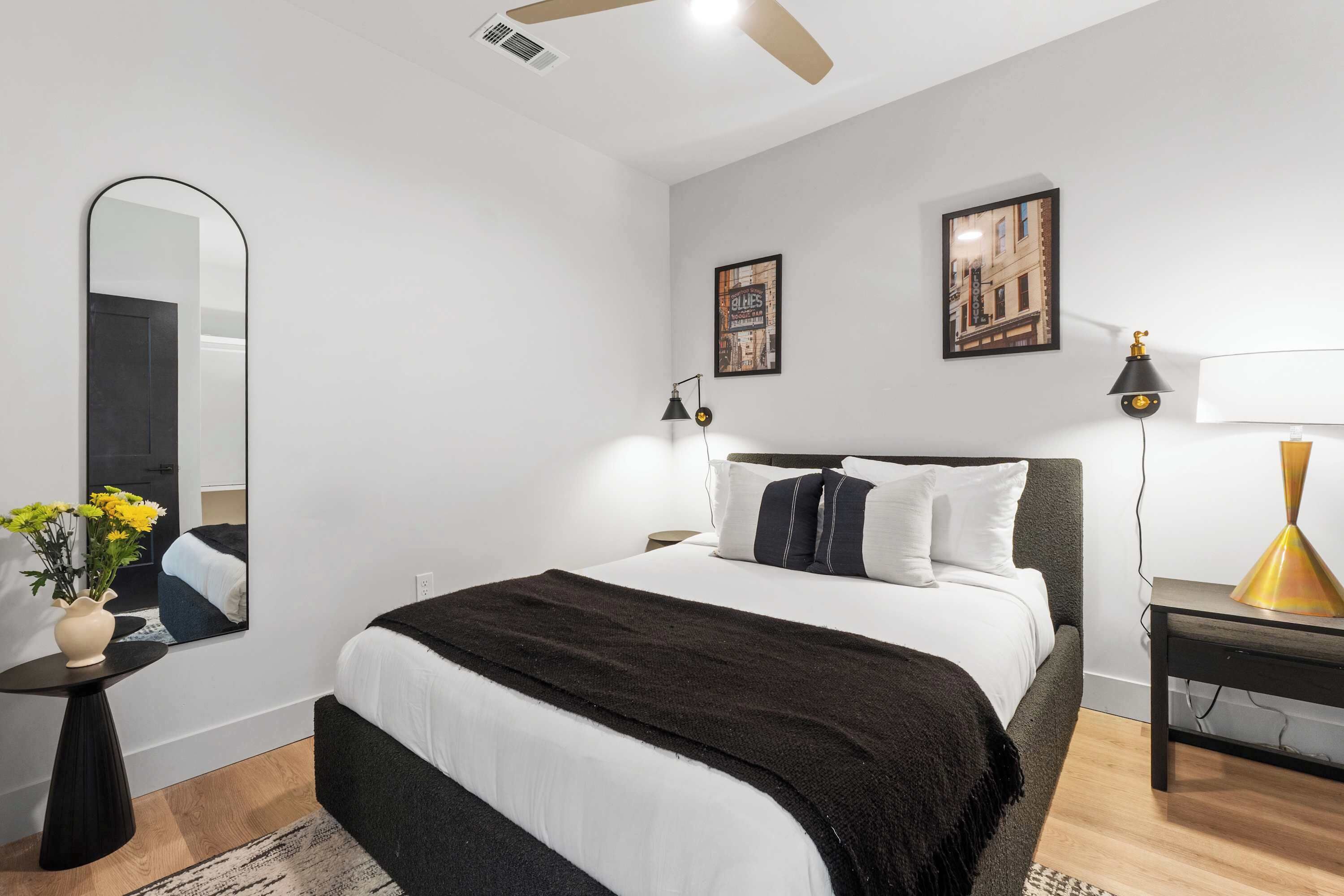 Un dormitorio moderno con cama, espejo, mesita de noche, lámpara y una pared con dos cuadros enmarcados.