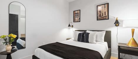 Un dormitorio moderno con cama, espejo, mesita de noche, lámpara y una pared con dos cuadros enmarcados.