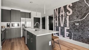 A modern kitchen with a central island, stainless steel appliances, and a black and white cityscape mural. - Urban & Soulful Escape: 2 Condos Near Vandy | Pool (Nashville)
