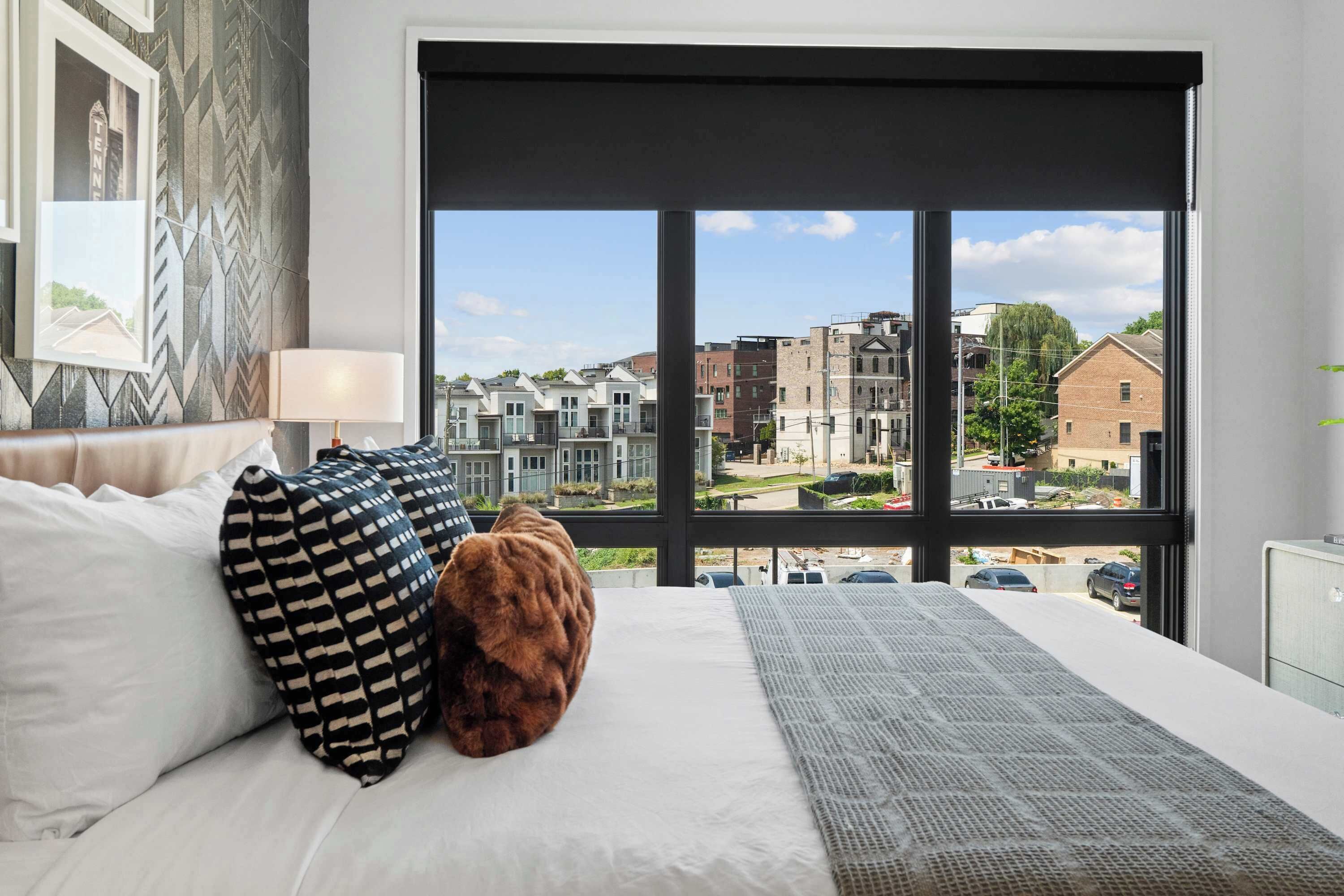 A bedroom with a large window, a bed with white bedding, patterned pillows, and a brown throw.