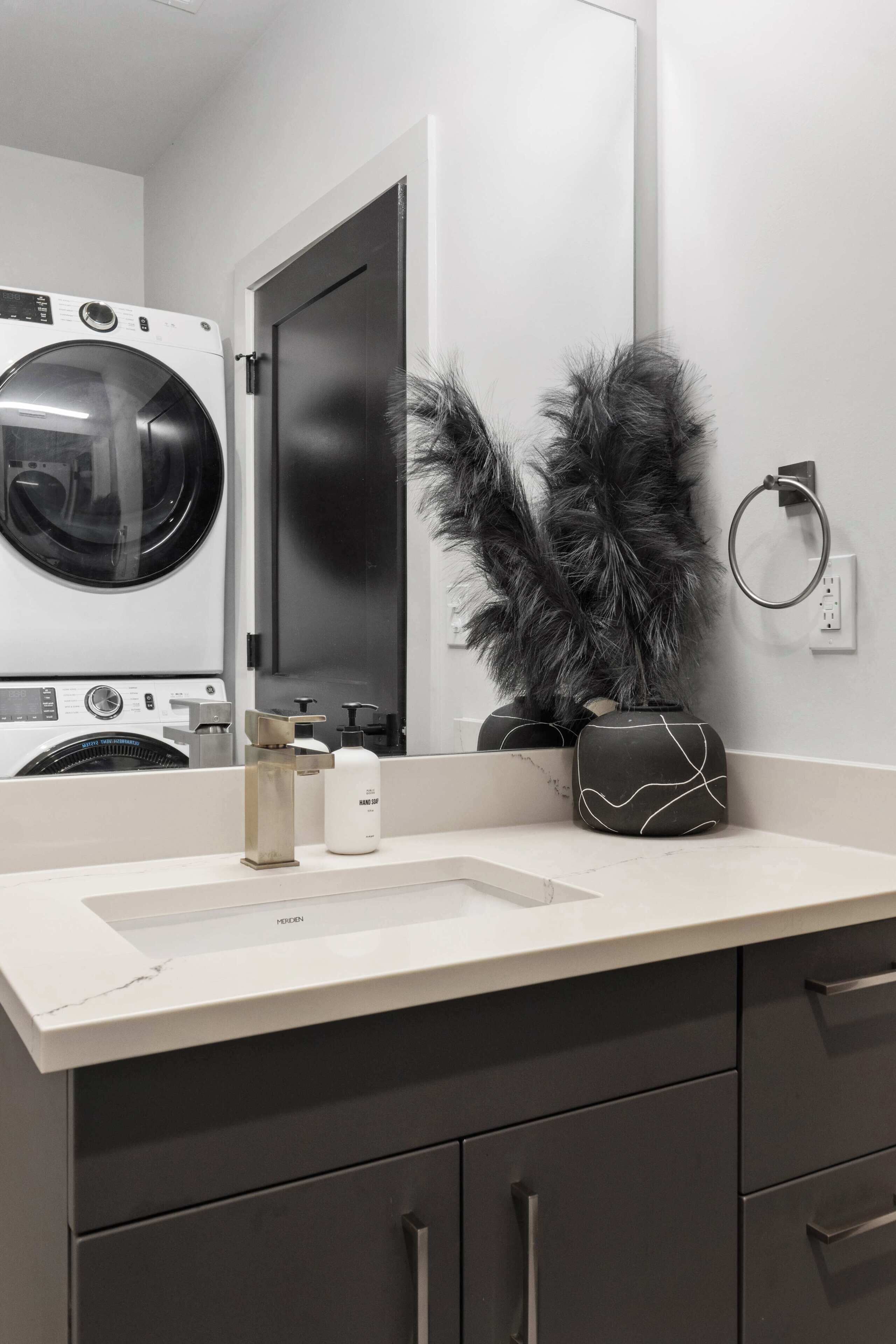 Un baño moderno con un lavamanos blanco, un espejo, una lavadora y un perchero para toallas.