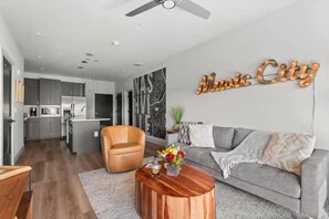 A modern living room with a grey sofa, a wooden coffee table, and a kitchen in the background. - Urban & Soulful Escape: 2 Condos Near Vandy | Pool (Nashville)