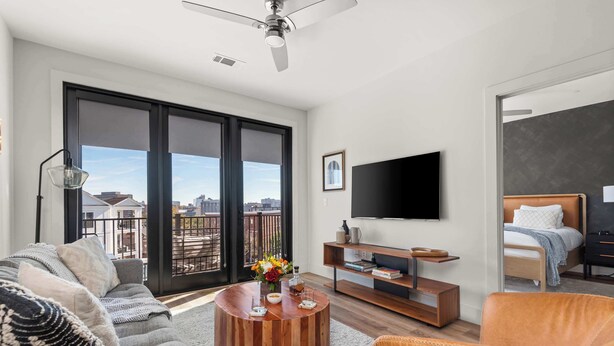 A modern living room with a sofa, coffee table, TV, and a balcony with a city view. - Urban & Soulful Escape: 2 Condos Near Vandy | Pool (Nashville)